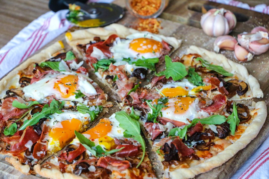 Breakfast pizza with arugula, soft bakes eggs, caramelized onions, and prosciutto